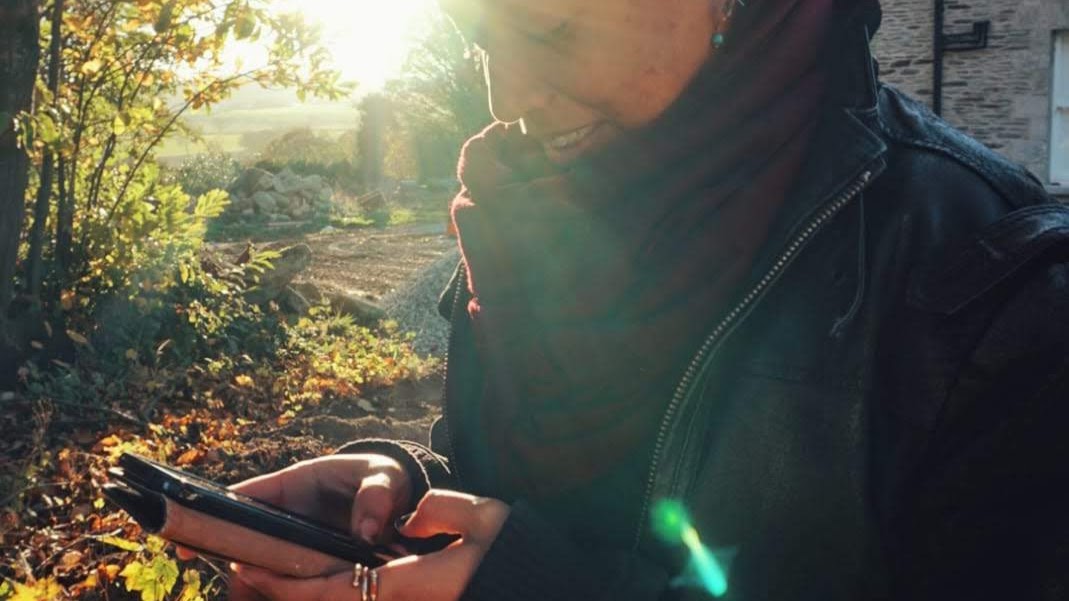 Brittany is smiling, holding and staring at her phone. She's wearing a coat and a scarf and standing outdoors.