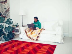 A woman is sitting on a white sofa with a blanket and laptop on her lap. The woman is wearing a green jumper and staring at the laptop screen