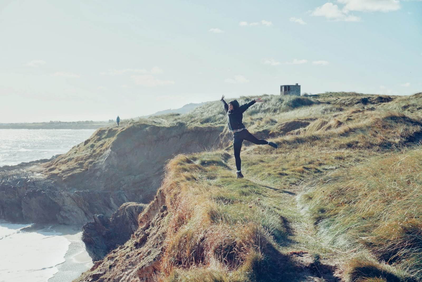 woman in dark clothes jumping into the air near the edge of a grassy cliff