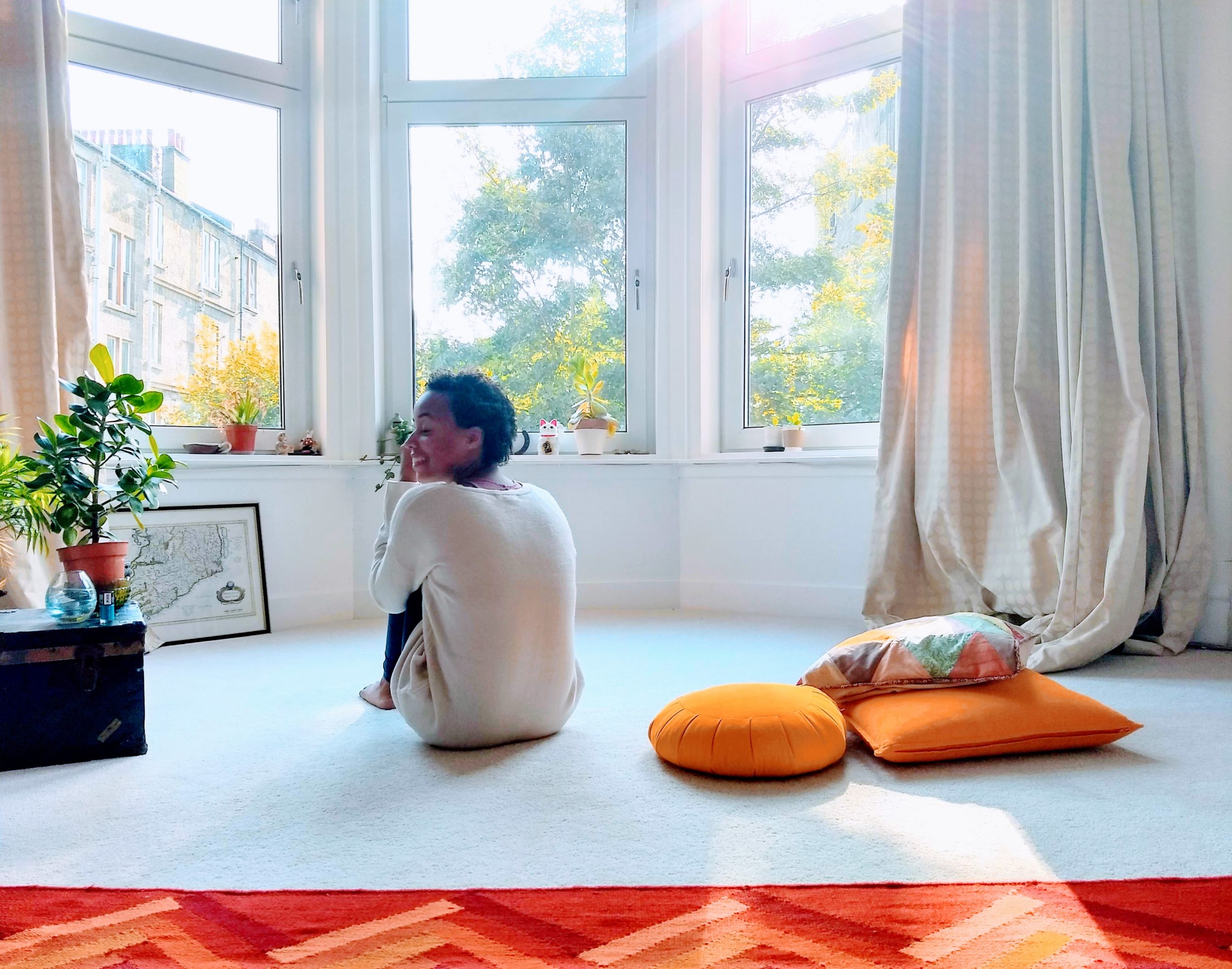 Brittany, a woman and a Bahamian with Multiple Sclerosis is sitting on a white carpeted floor in front of a sunny bay window in an oversized white jumper. Brittany's back is facing us, she is looking to the left and smiling over her shoulder