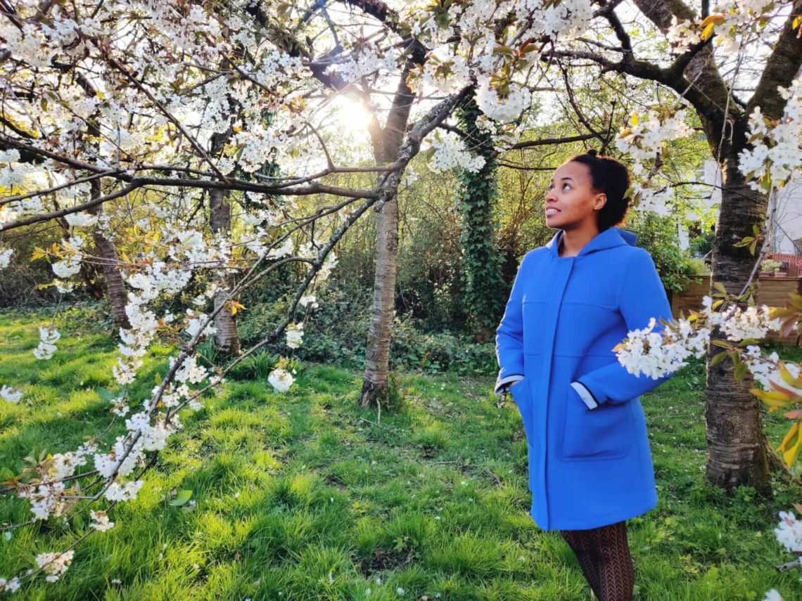 A woman in a bright blue coat is standing under a tree of cherry blossoms. She is facing the camera, smiling and looking to the left. She is travelling with ms