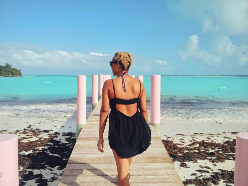 Brit's back in a black backless sun dress at the treasure cay public dock. The beach is flanking the dock and crystal blue water is in the background.