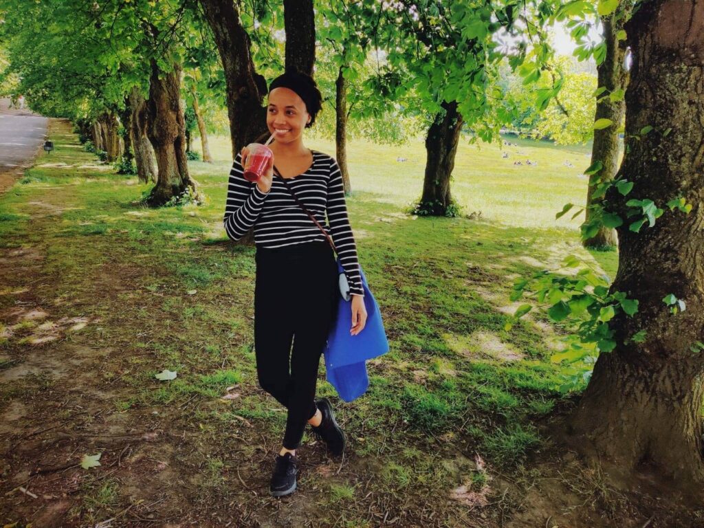 A woman is posing in a black and white striped top and black jeans whilst enjoying a smoothie in the park and managing travelling with MS