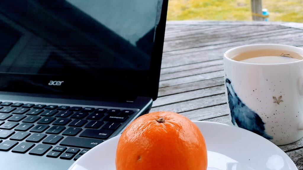 Acer laptop, orange on a plate and a cup of tea on a patio table outside as I cope with stress of unemployment during COVID