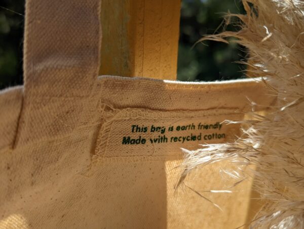 A close up of the inner tag of the tote bag which states, " This bag is earth friendly >> Made with recycled cotton." There is pampas grass on the side of the label