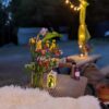 There are multi-coloured flowers in a vase on a metal picnic table next to a sheepskin throw. There are fairy lights above it and bags and jackets in a pile on a table in the background. It's dusk and there is a building structure in the background amongst dense woods.