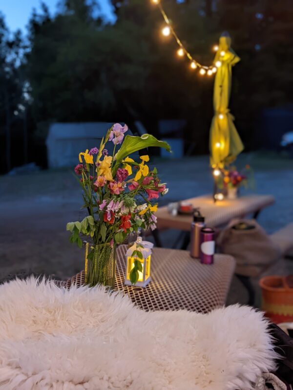 There are multi-coloured flowers in a vase on a metal picnic table next to a sheepskin throw. There are fairy lights above it and bags and jackets in a pile on a table in the background. It's dusk and there is a building structure in the background amongst dense woods.