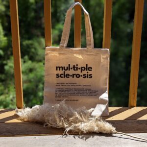 A canvas tote bag with the definition of Multiple Sclerosis printed in black on one side in the centre is propped up against a wooden railing on a balcony. There is a piece of pampas grass on the balcony in front of the bag. There are dense trees in the background and it's a sunny day.