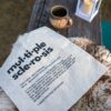 A canvas tote bag with the definition of Multiple Sclerosis printed in black on one side in the centre is lying on top of a sheepskin on a wooden bench next to a mug and a white lantern