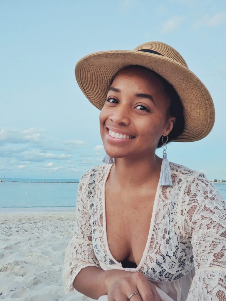 Brit is smiling, wearing a pink dress with lace at the top and on the sleeves. She is also wearing a straw hat. The beach is in the background.