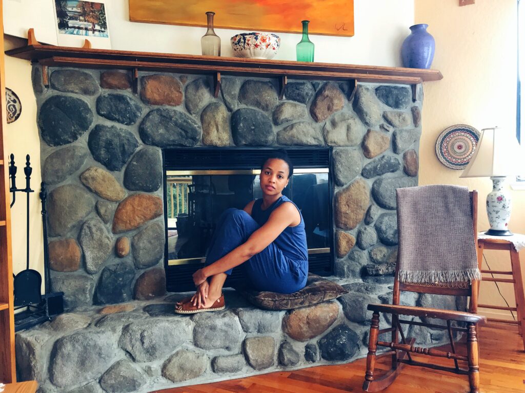 Brit is wearing a blue jumpsuit and sitting by a stone fireplace