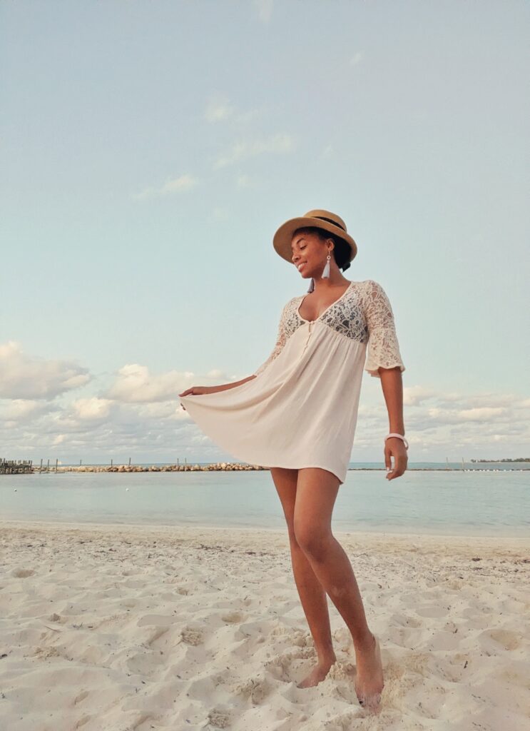 Brit is wearing a straw hat and a light pink flowy, short dress with a lace top. The beach is in the background.